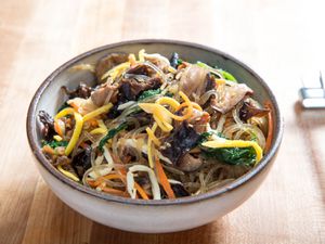 A bowl of jap chae showing colorful egg strips, spinach, carrot, and more intertwined in the noodles.