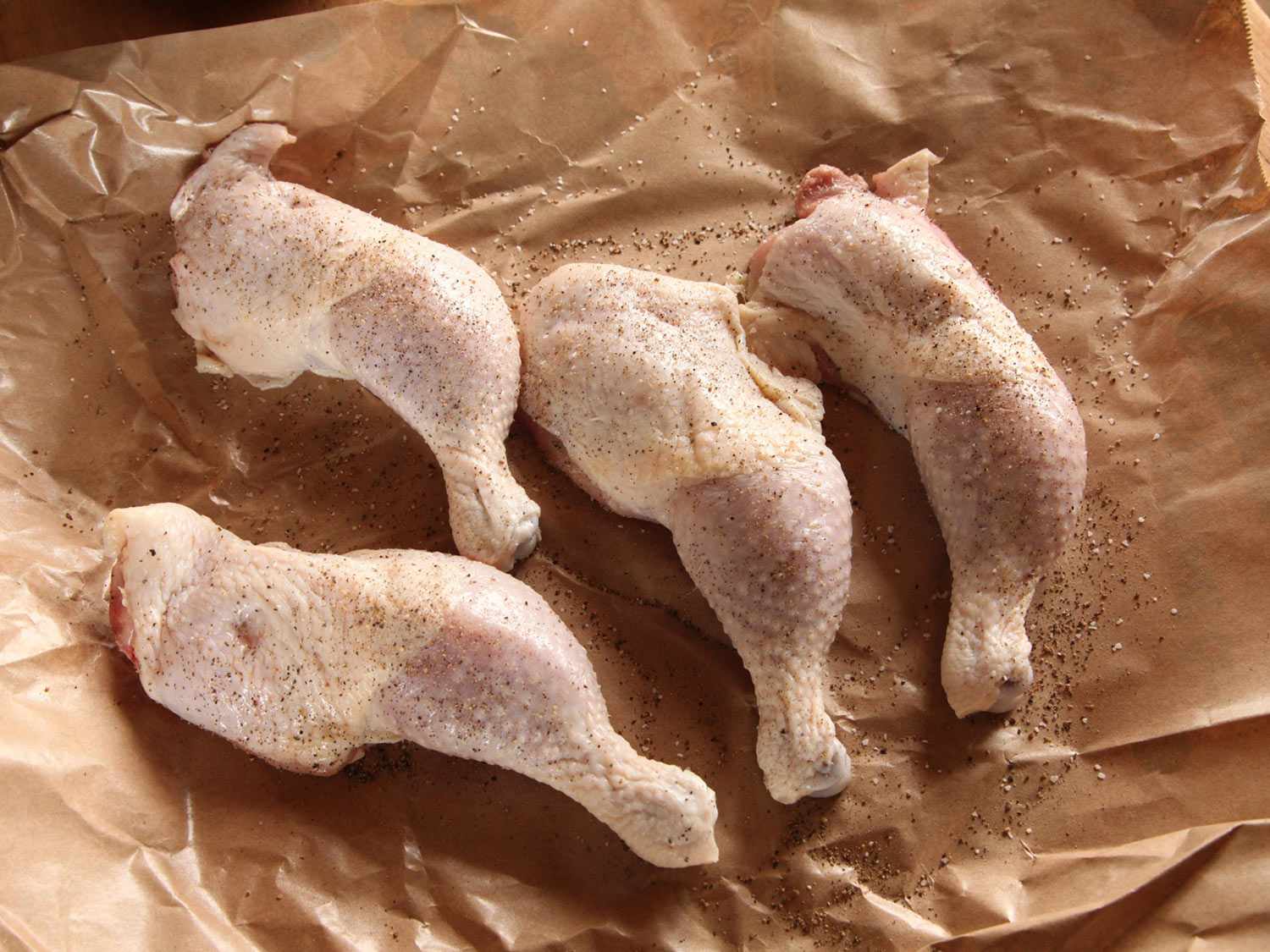 Four chicken legs on a crinkled piece of brown wax paper, sprinkled with black pepper.