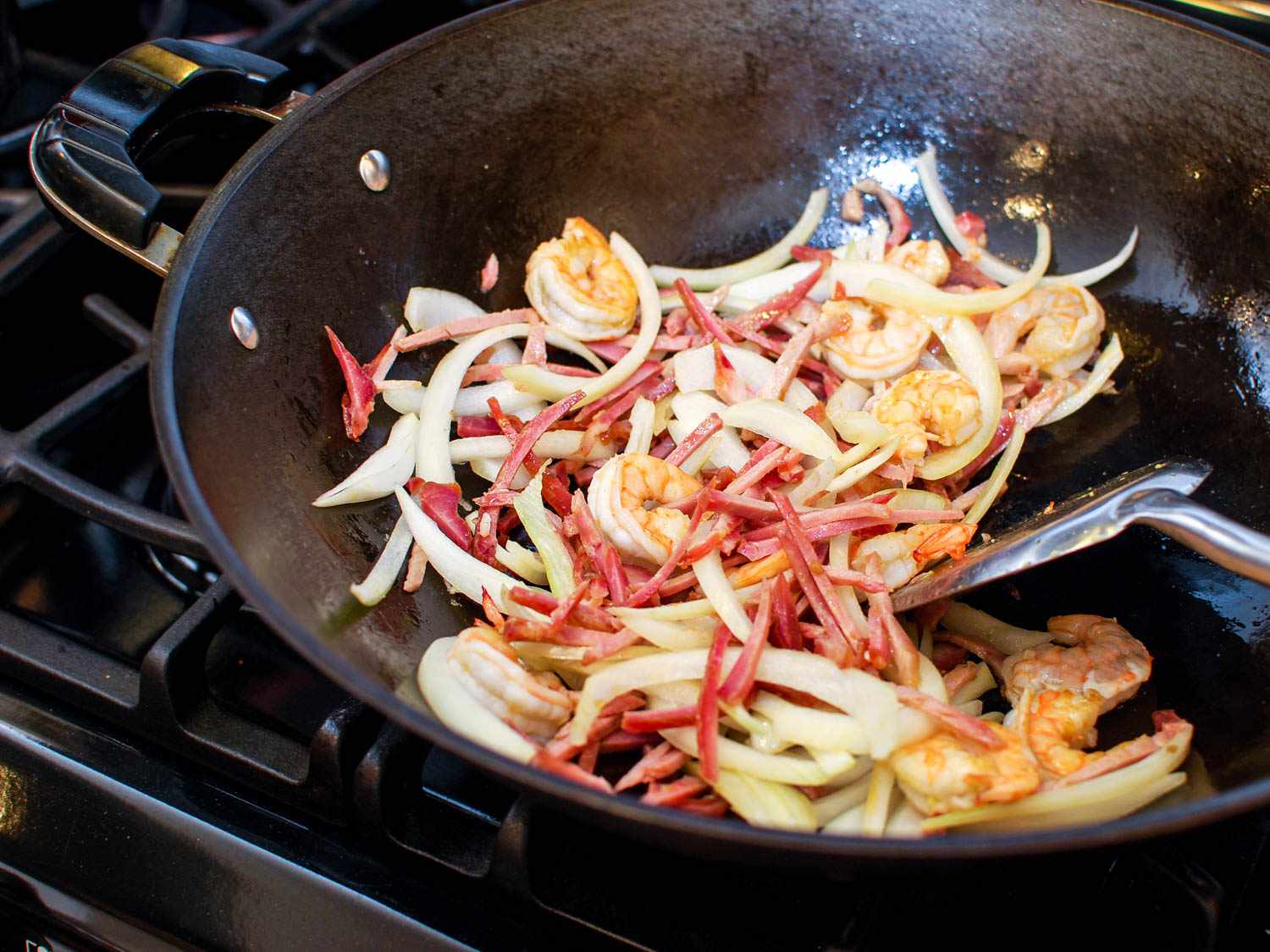 Spatula stirring shrimp, onions, snow peas, carrots, and char siu in a hot wok.