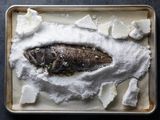 被埋在烤盘上的盐堆里的一整条烤黑鲈鱼，在盐壳破裂后被取出，露出了水面。