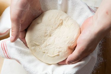两只手正在把一团面包面团放进碗里，碗里铺着撒了面粉的厨房毛巾。
