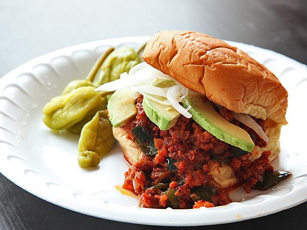 A chorizo Sloppy Joe sandwich topped with sliced avocado and thinly sliced onions on a white styrofoam plate. Some pickled peppers are on the side of the plate.