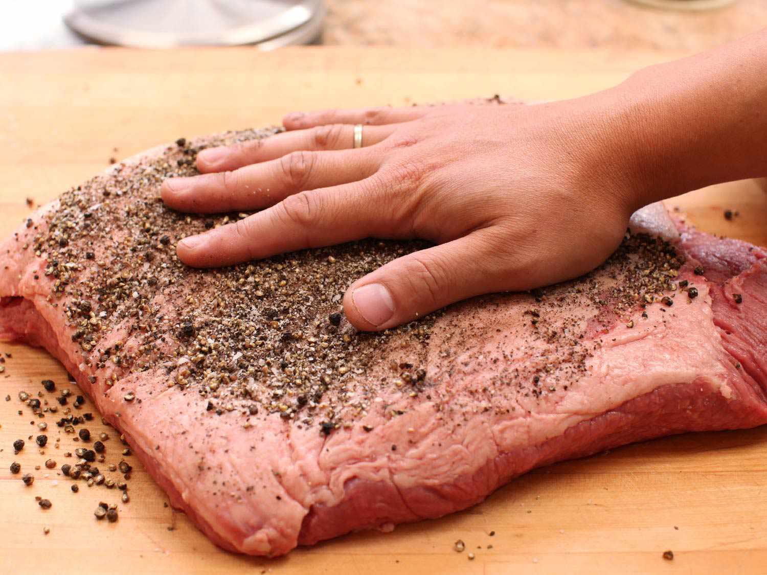 A hand rubbing coarsely ground peppercorns over a slab of brisket