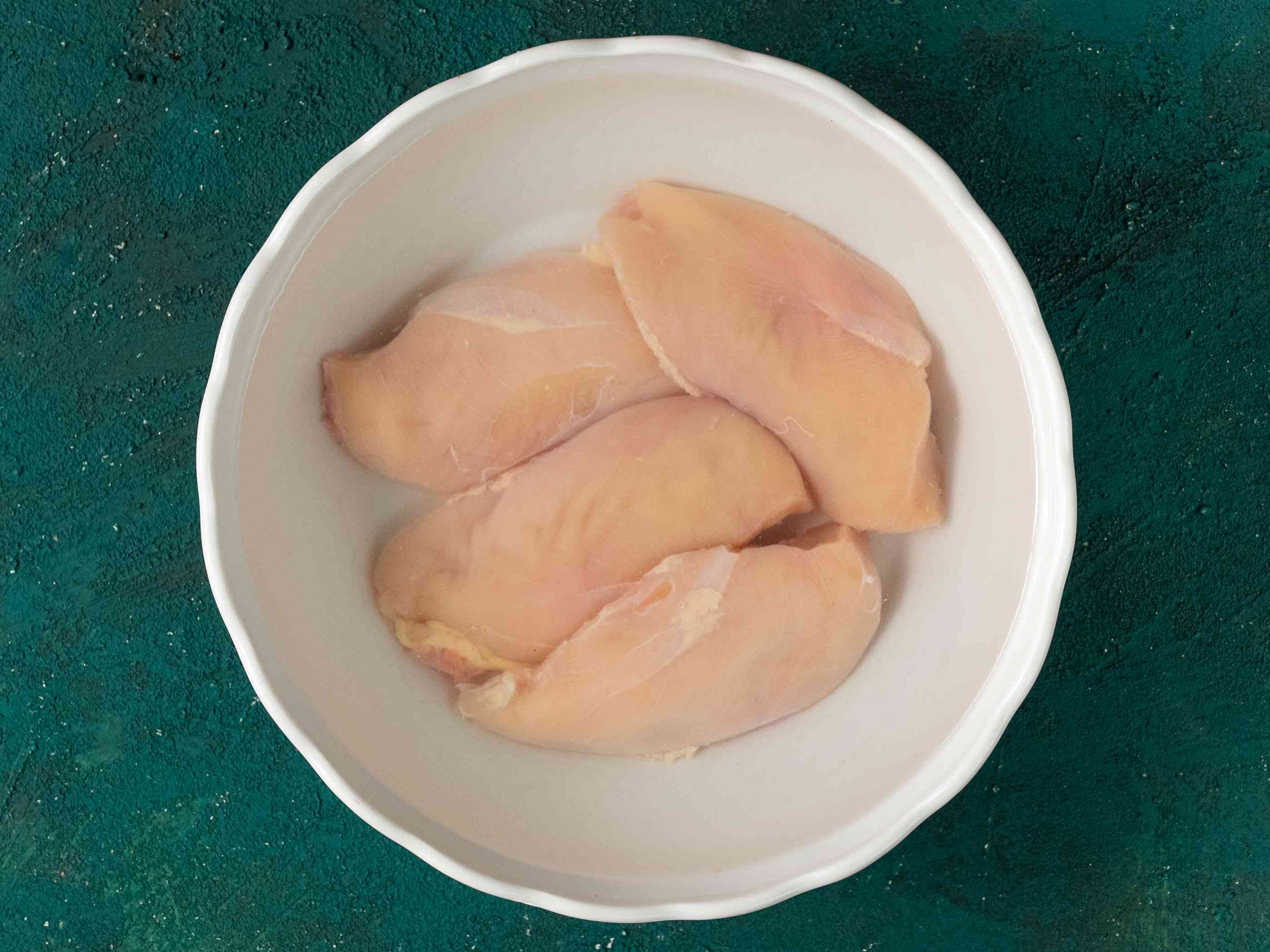 半块鸡胸肉浸在盐水里，放在一个白色陶瓷碗里，背景是蓝绿色。