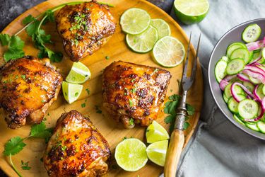 Four Vietnamese baked chicken thighs, served on a wooden serving platter with cut limes and a bowl of cucumber salad.