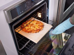 A hand with an oven mitt pulling a baking stone with a pizza on it out of the oven