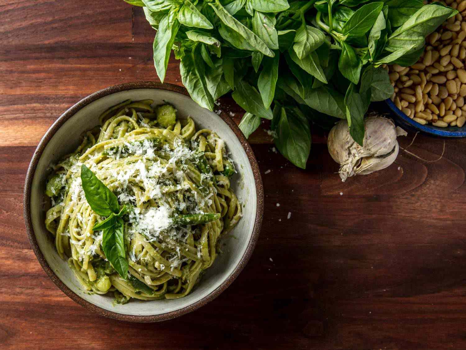 Pesto on linguine in a bowl on rustic tabletop, with fresh basil, garlic, and pine nuts on the side.