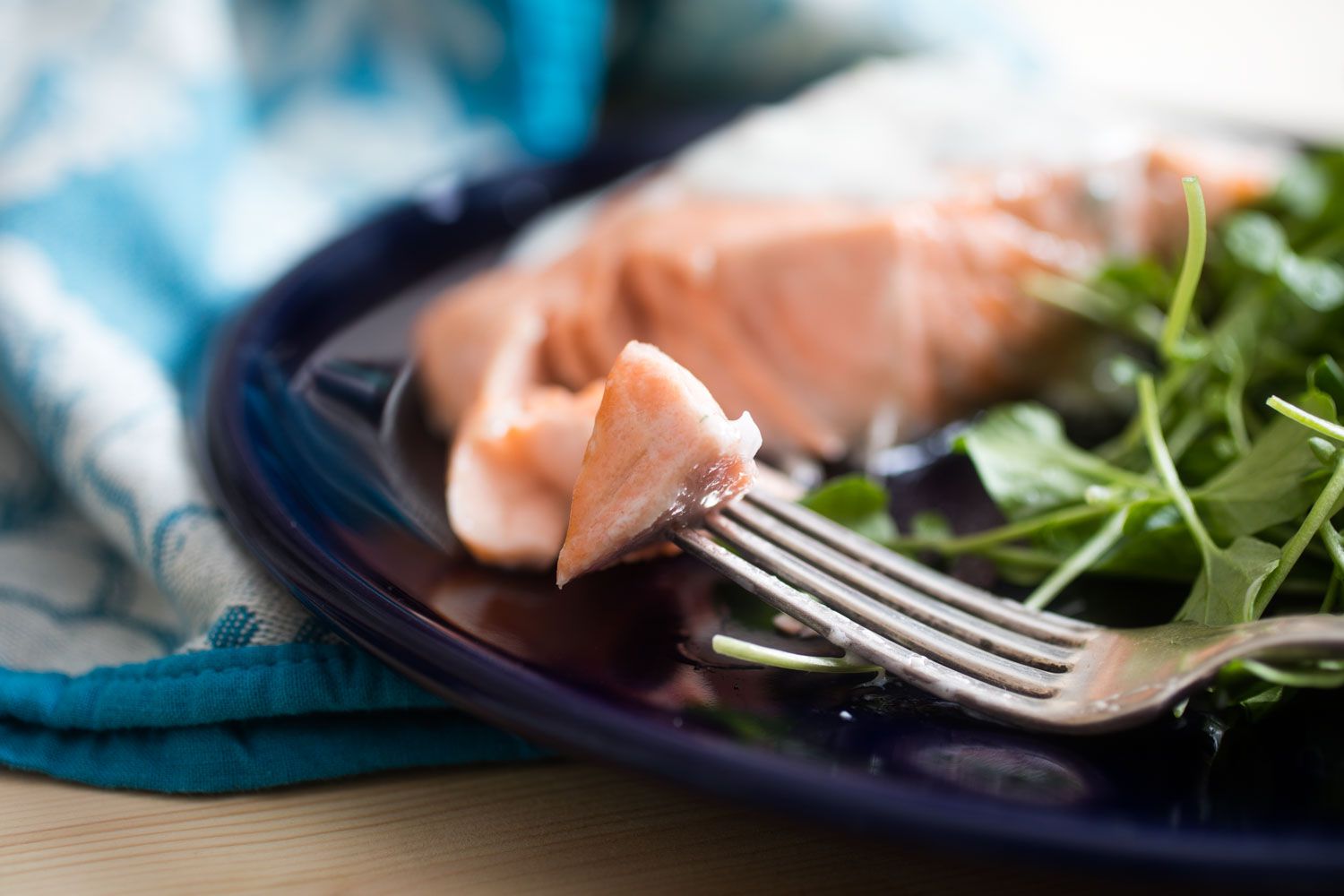 把三文鱼串在叉子上咬一口。叉子放在一个深蓝色的盘子里。在模糊的背景中有更多的鲑鱼和绿色蔬菜。gydF4y2Ba