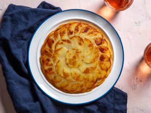 A golden brown pommes anna sits on a serving plate, showing beautiful shingled rounds of thinly sliced potato.