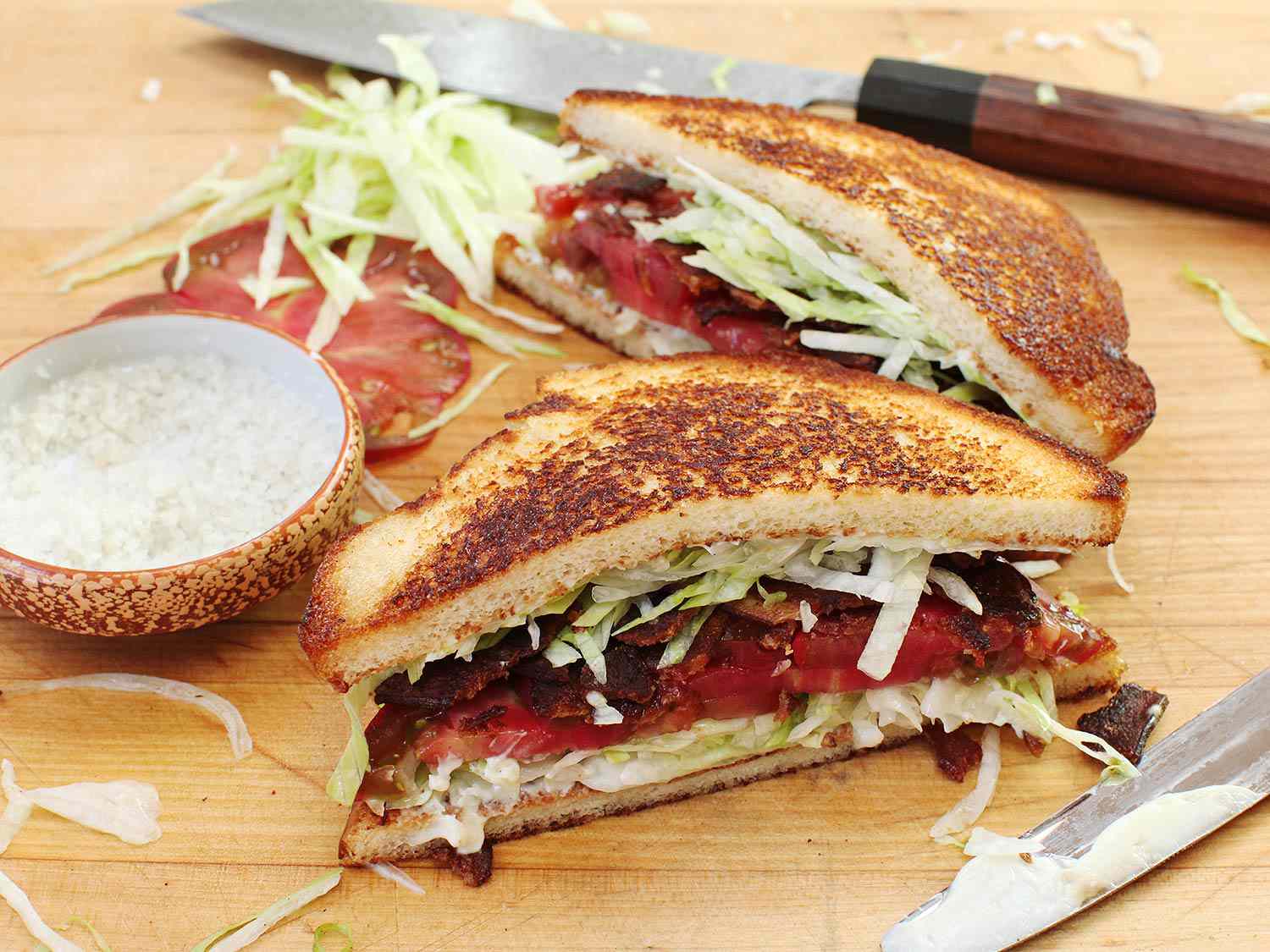 将BLT三明治对角切成两半，旁边放一小碗粗盐，番茄切片和生菜丝
