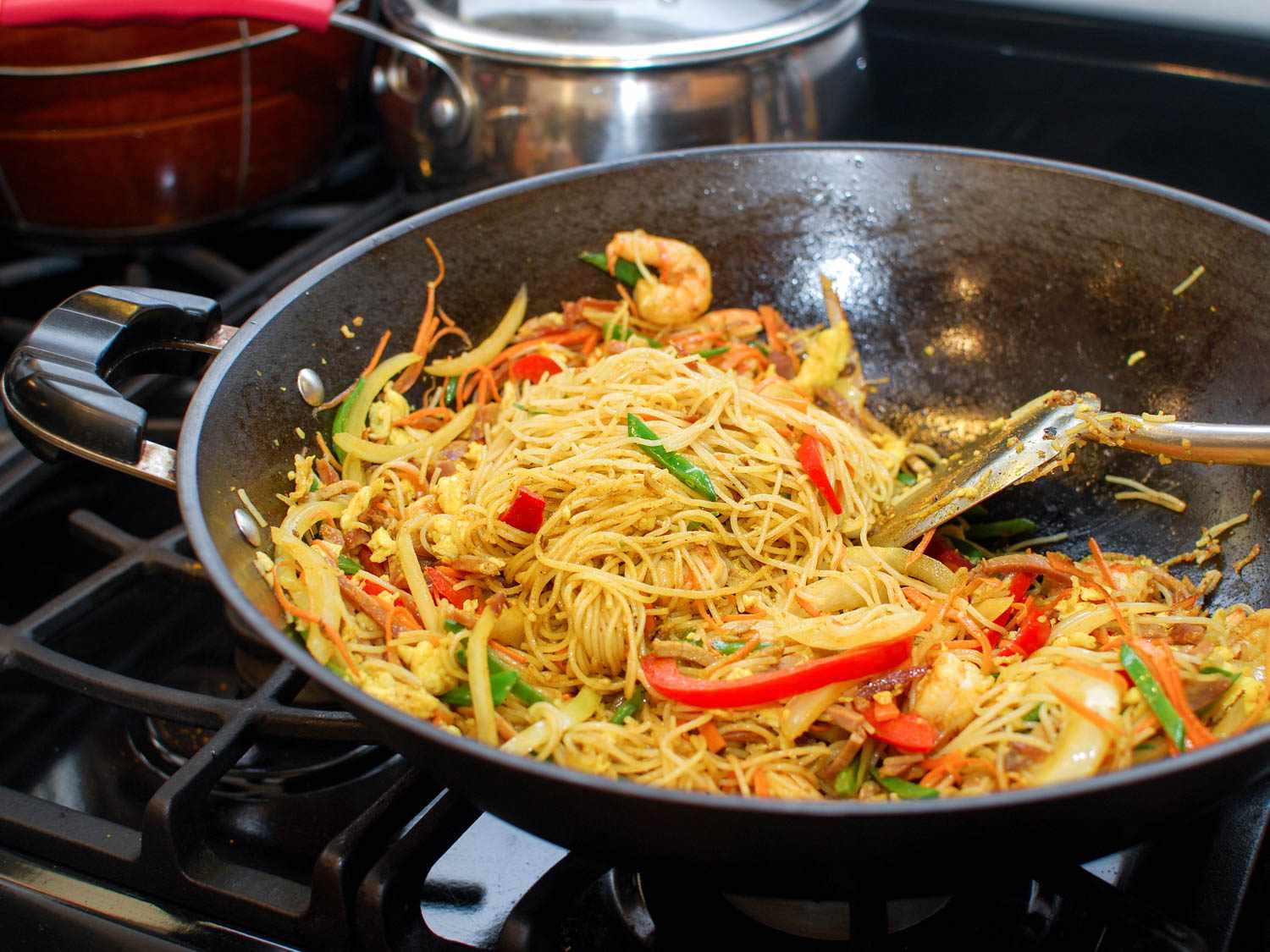 Spatula combining egg, meat, vegetables, and curried vermicelli noodles in a hot wok.