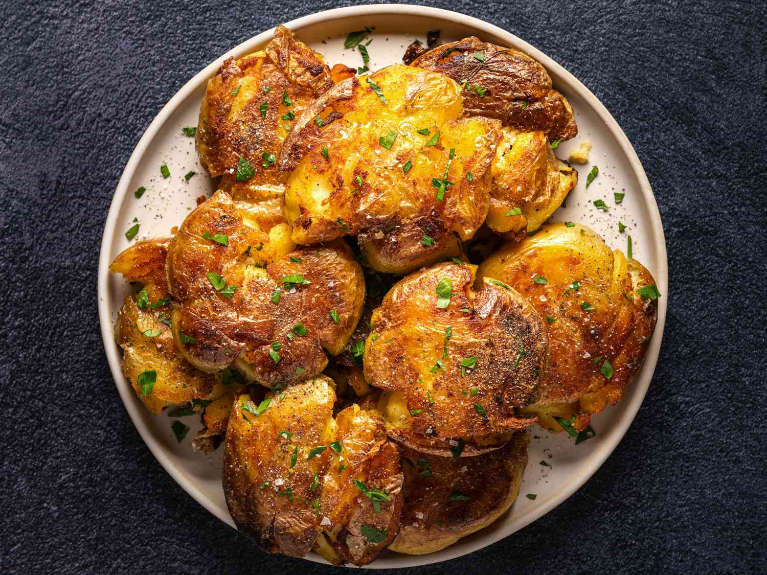 Golden brown crispy smashed potatoes on an oval white plate on a dark textured background.