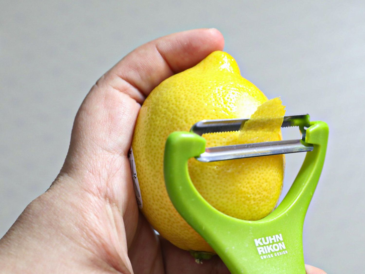 A hand holding a lemon as a y-peeler peels a strip of lemon peel