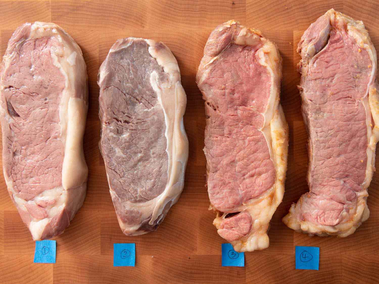 俯视四条带牛排烹饪真空，由编号胶带在砧板上识别。左边的第二块牛排比其他三块要灰白得多。