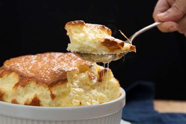 A large savory cheese souffle with someone holding a spoon of souffle above the casserole.