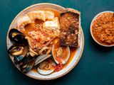 一份盛在碗里的cioppino，盛满了饱满的虾、贻贝、蛤蜊、鱿鱼、鱼等。旁边有一片烤得很深的酵母，还有一个小碗，里面放着烤红辣椒调味品。