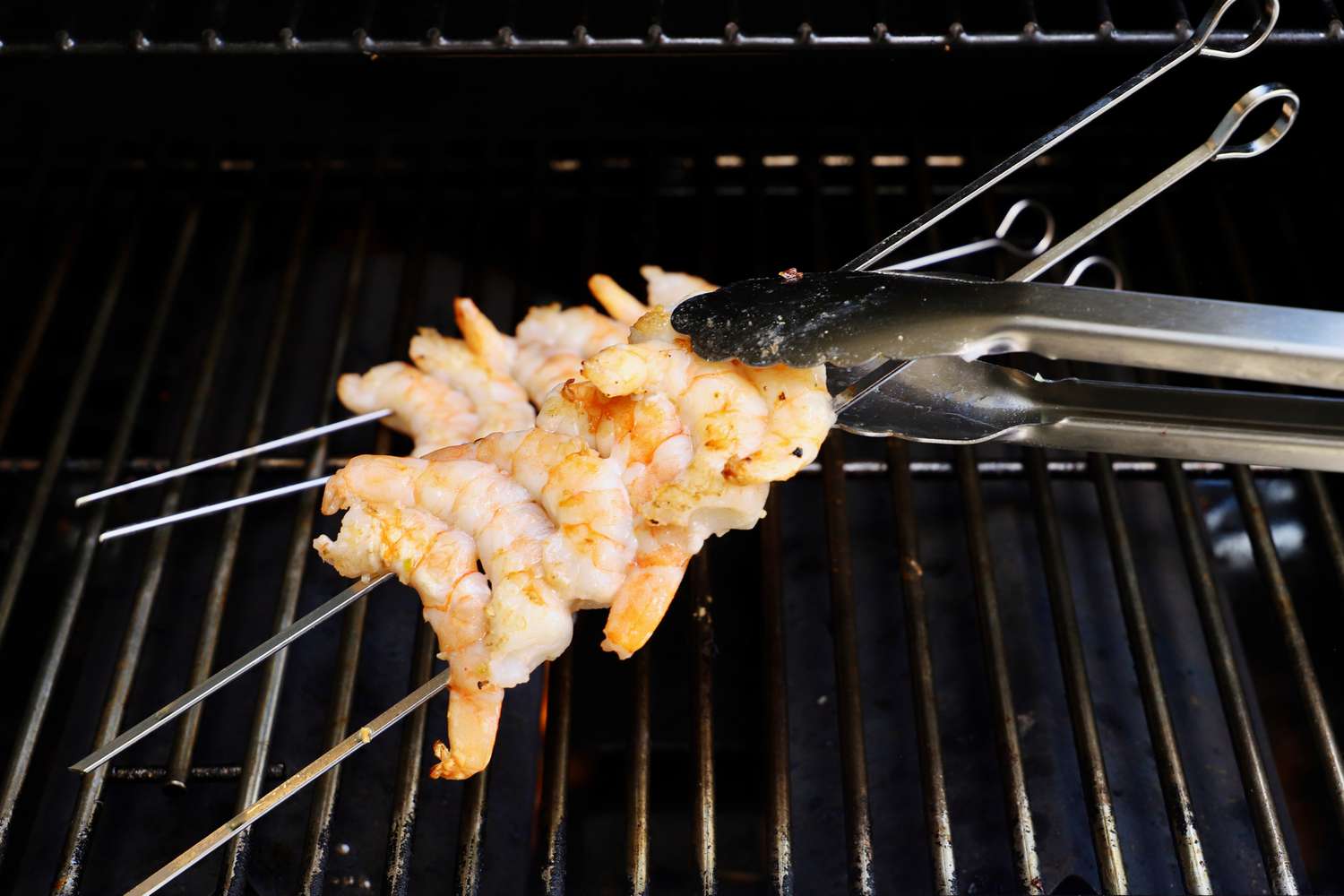 a closeup look at tongs flipping two skewers that have shrimp threaded onto them