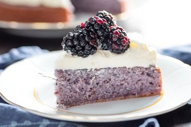 Profile view of a slice of blackberry cake with cream cheese frosting, served on a white plate and garnished with several large blackberries.