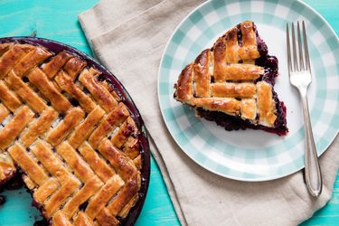 A slice of blueberry pie with a lattice crust next to the whole pie