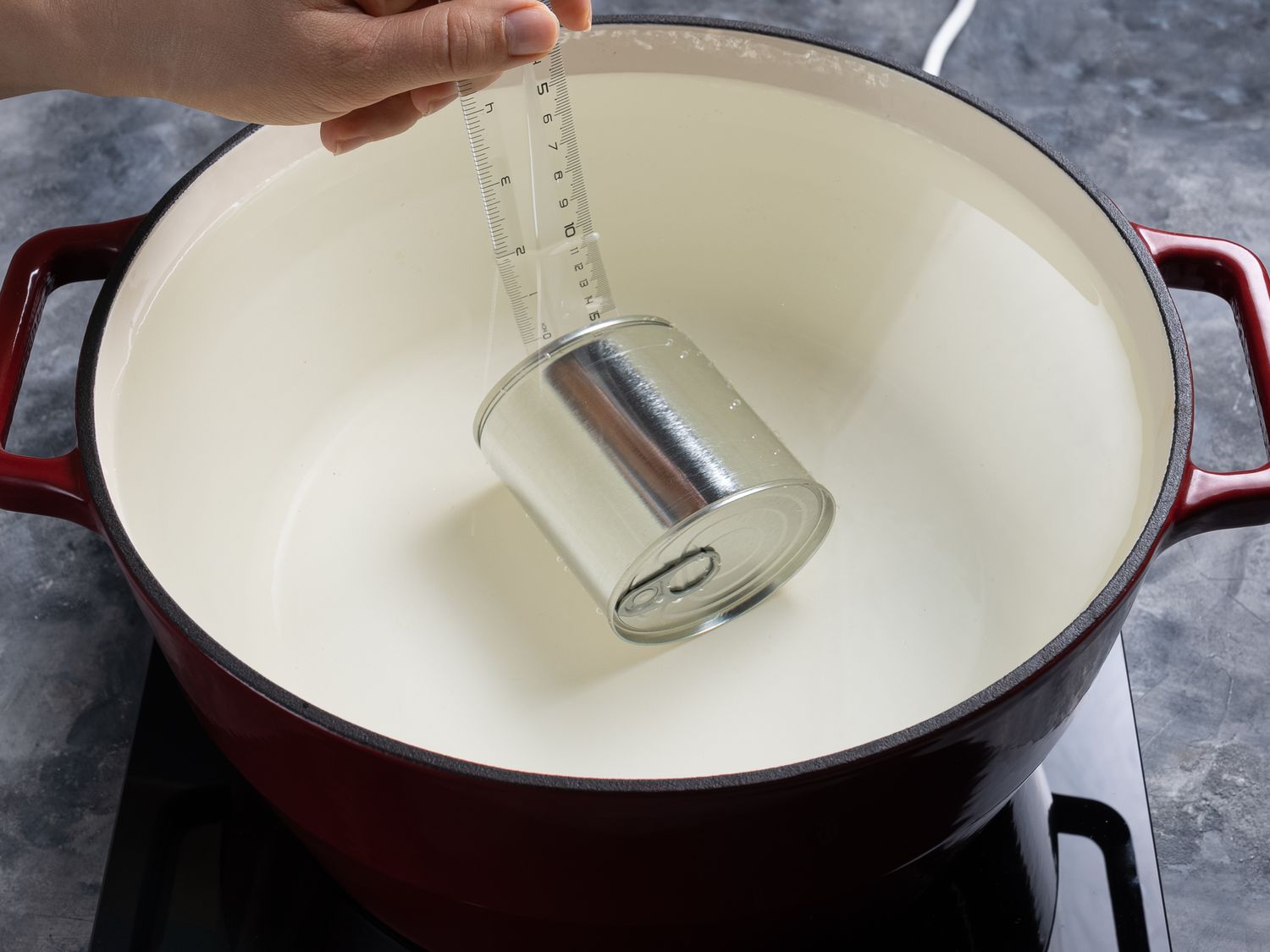 The can of condensed milk in a dutch oven filled with water, with a clear ruler placed in the pot to indicate how much water is covering the can