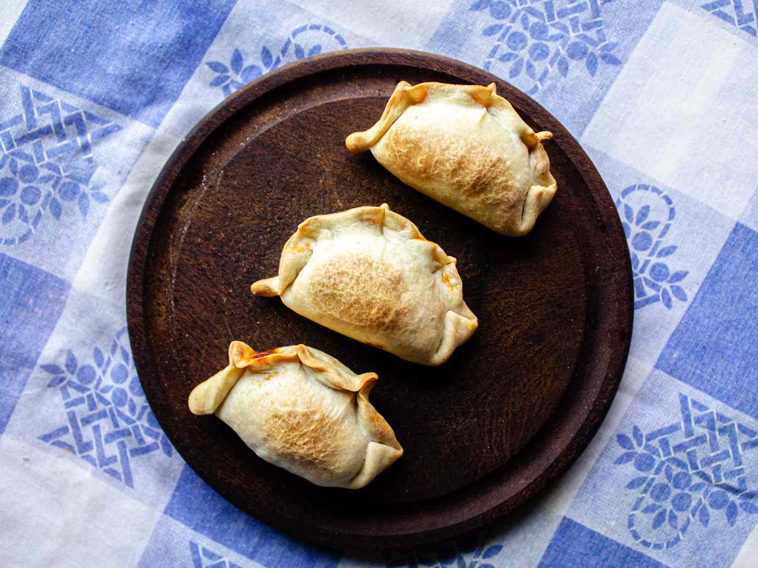 3个肉馅卷饼放在木盘上，上面是一种传统的阿根廷织物，有蓝白格子图案
