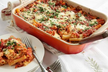 A casserole dish of baked ziti on a table. Next to the casserole is a stack of white plates. Some of the baked ziti has been scooped on the top plate.