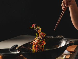 在黑暗的厨房里，一个人拿着筷子扔锅，导致五颜六色的食物悬浮在空气中