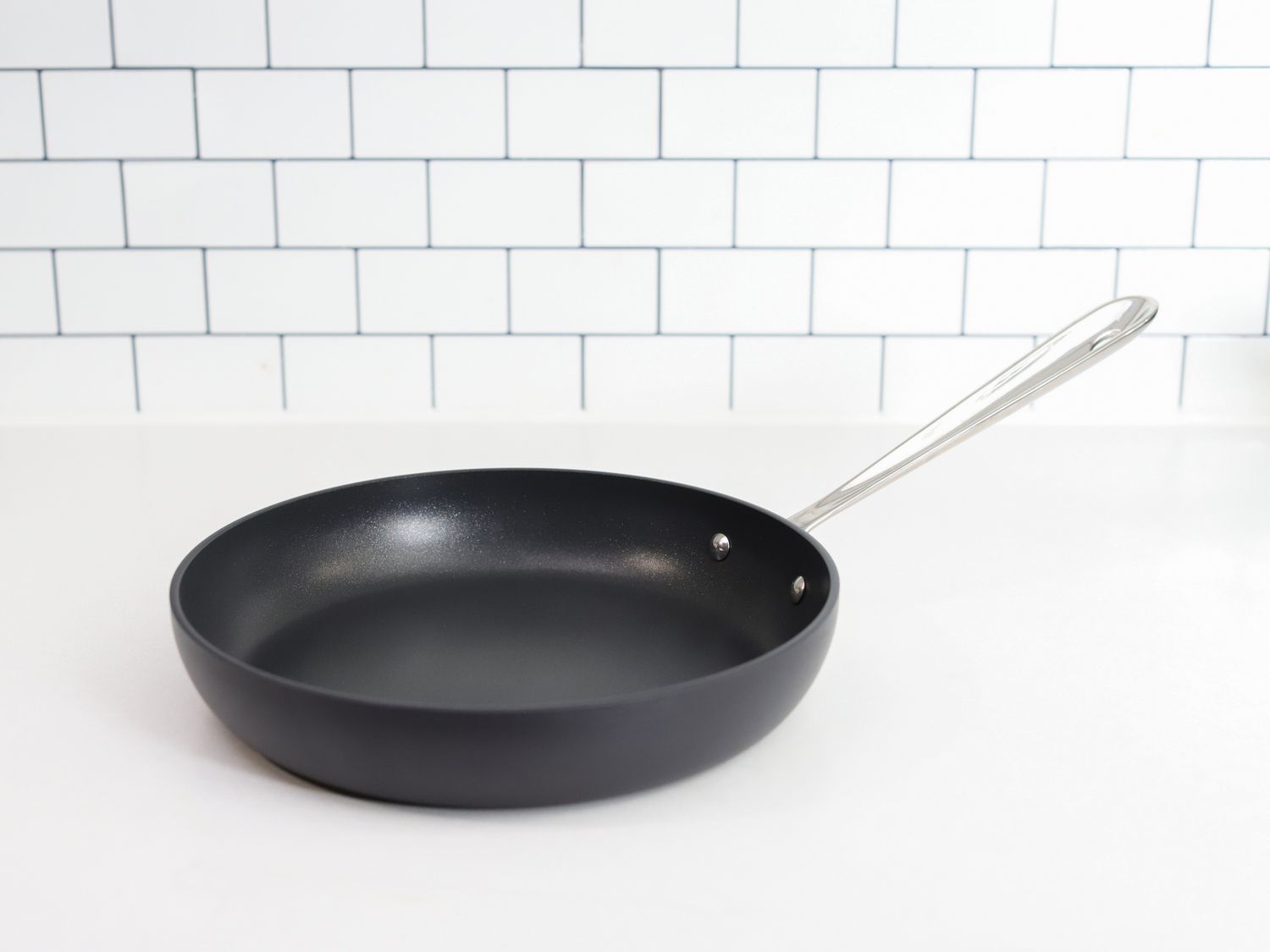 the all-clad nonstick skillet sitting on a white countertop with a white subway tile background behind it