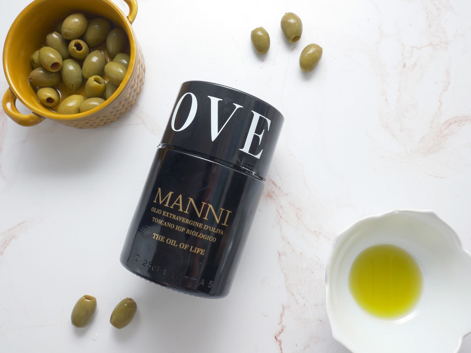 manni olive oil on a pale pink marble surface with olives and olive oil in a cup