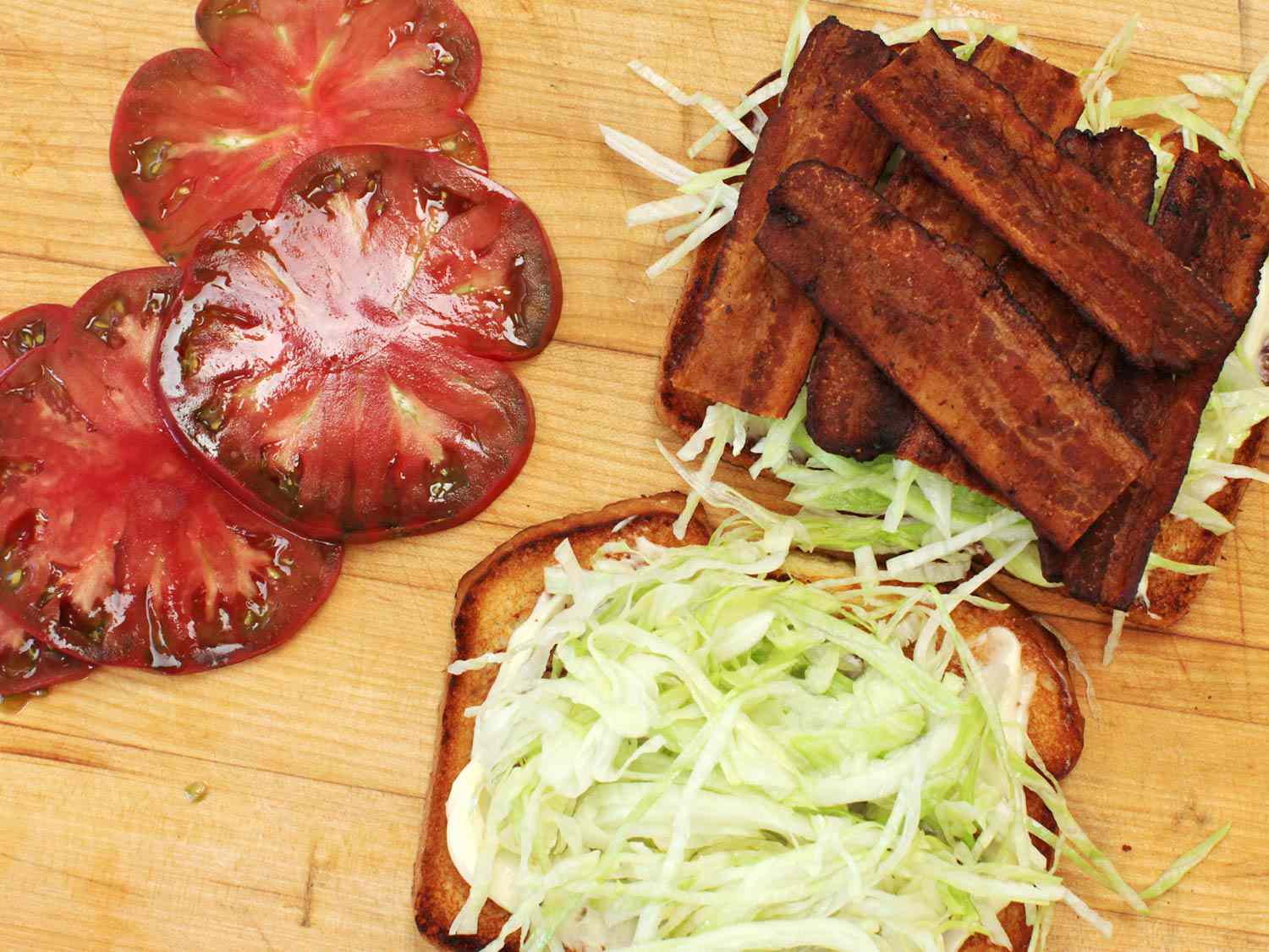 制作BLT三明治:将切片番茄放在一片涂有蛋黄酱和生菜丝的烤面包片旁边，另一片面包片上分层着培根和生菜