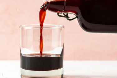 A bottle of DIY cherry liqueur on the right hand side of the image pouring liqueur into a short rocks cocktail glass on the left hand side of the image.