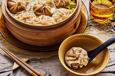 Angled view of a tablescape with a bamboo steamer full of dumplings behind a biten into soup dumpling on a soup spoon in an individual bowl on a black and white marbled surface. Overall tone and lighting is warm