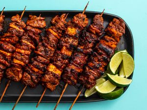A black platter of charred al pastor skewers and a pile of lime wedges for last-minute drizzling.