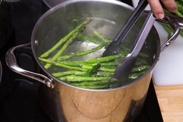 Asparagus being blanched in a large stock pot filled with water. Someone is removing the asparagus with a pair of tongs.