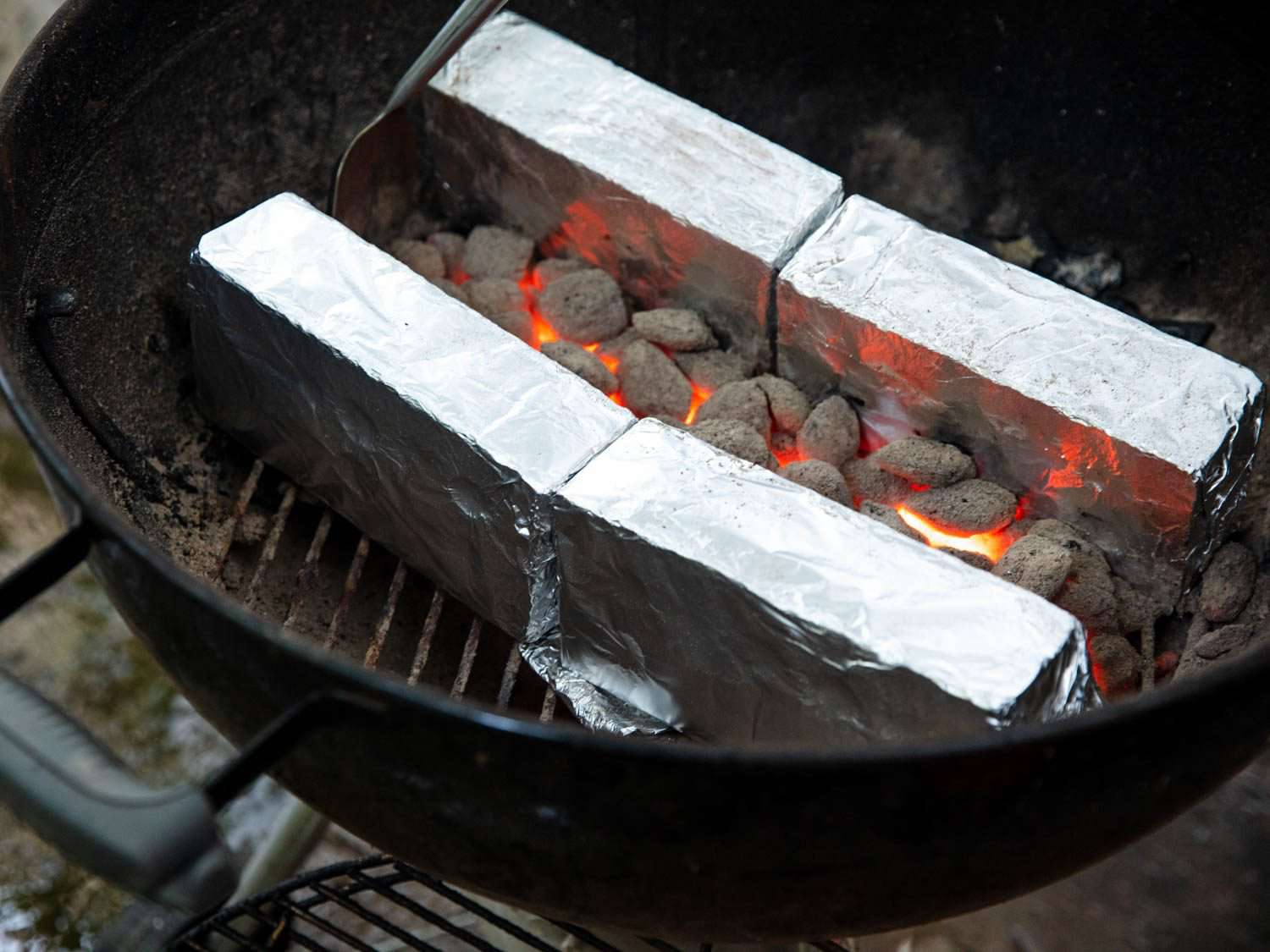 Arranging glowing coals between two parallel walls of foil-wrapped bricks for cooking skewers in a kettle grill.