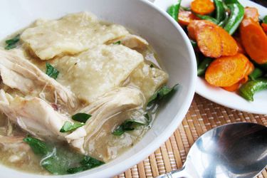 A soup bowl with a helping of chicken and slick dumplings. Sautéed carrot slices and snap peas are piled on a plate nearby.
