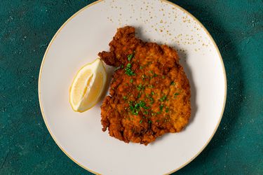 A golden brown fried chicken schnitzel cutlet with a lemon wedge on a gold and white ceramic plate on a teal-colored textured background.