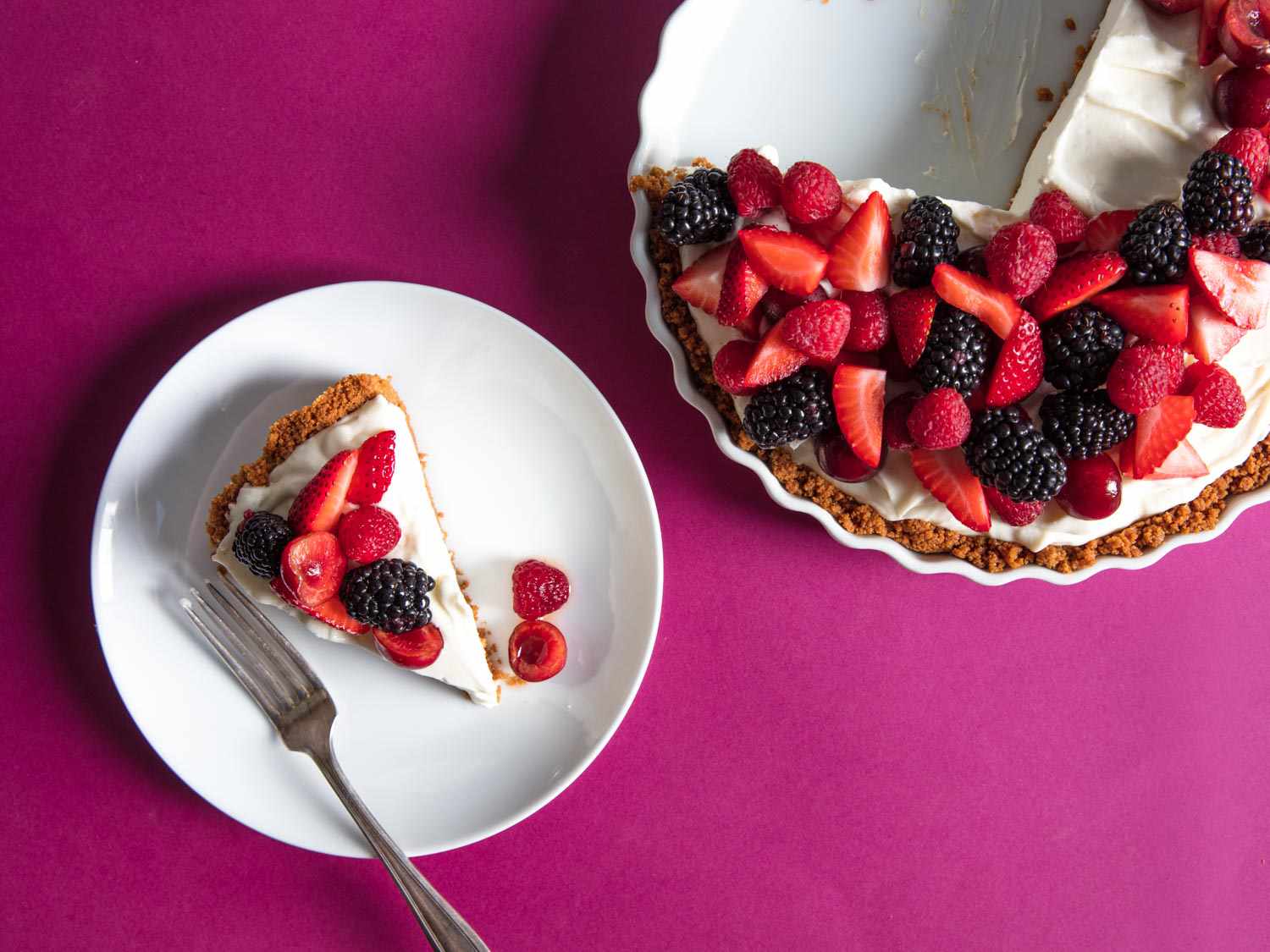 头顶上点缀着浆果的芝士蛋糕和一块放在品红色台布上的镀金蛋糕。＂width=