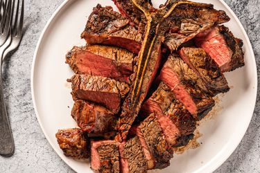 A t-bone (porterhouse) steak on a white plate, with the sliced meat placed around the bone. On the lefthand side of the image are several metal forks stacked on top of each other.