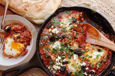 Shakshuka in a cast iron skillet, topped with parsley and olives, with a scooped single serving in a bowl and pita bread on the side.