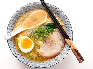 一碗tonkatsu拉面，上面放半个溏心蛋、葱和一片猪肉。碗边上放着一把中国汤勺和一双筷子。