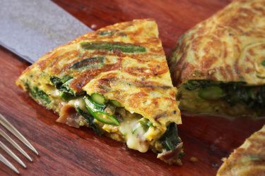 Closeup of a slice of flipped frittata with asparagus, spinach, ham, and cheese bering lifted from a cutting board with a chef's knife.