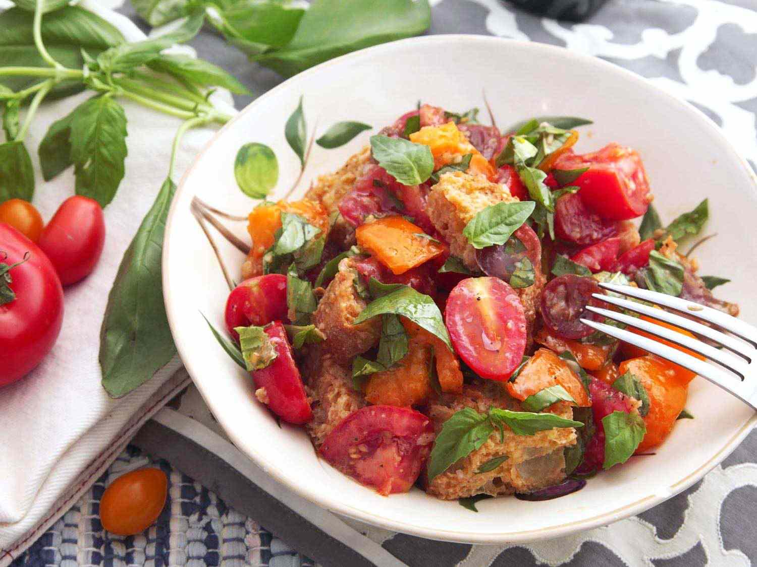 Close-up of finished panzanella (tomato and bread salad) in a white dish, next to ripe tomatoes and basil sprigs