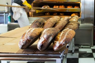 塔廷面包店厨房里面包师的桌子上放着几条面包。背景中，有人正在用一根长柄皮在商用烤箱中移动面包。