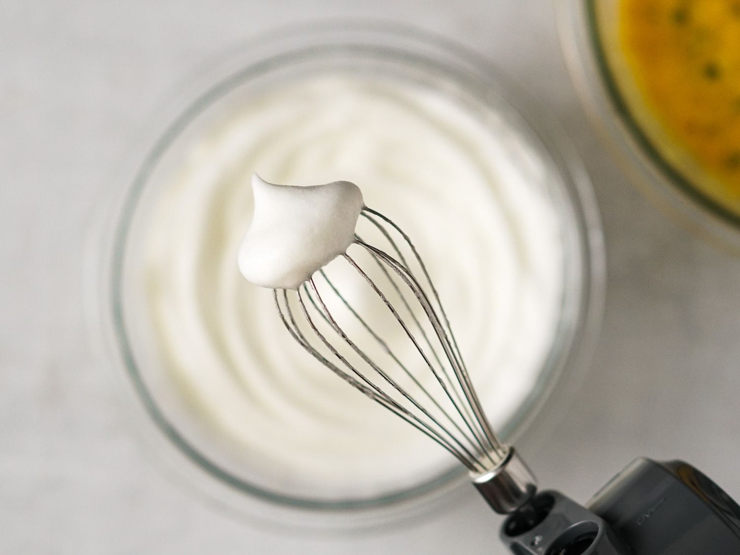 A bowl of whipped egg whites, with a beater held up to the camera displaying the texture of the fluffy egg whites.