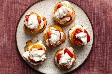 A top-down image of six composed strawberry shortcakes, on a speckled ceramic plate on a textured cloth background.