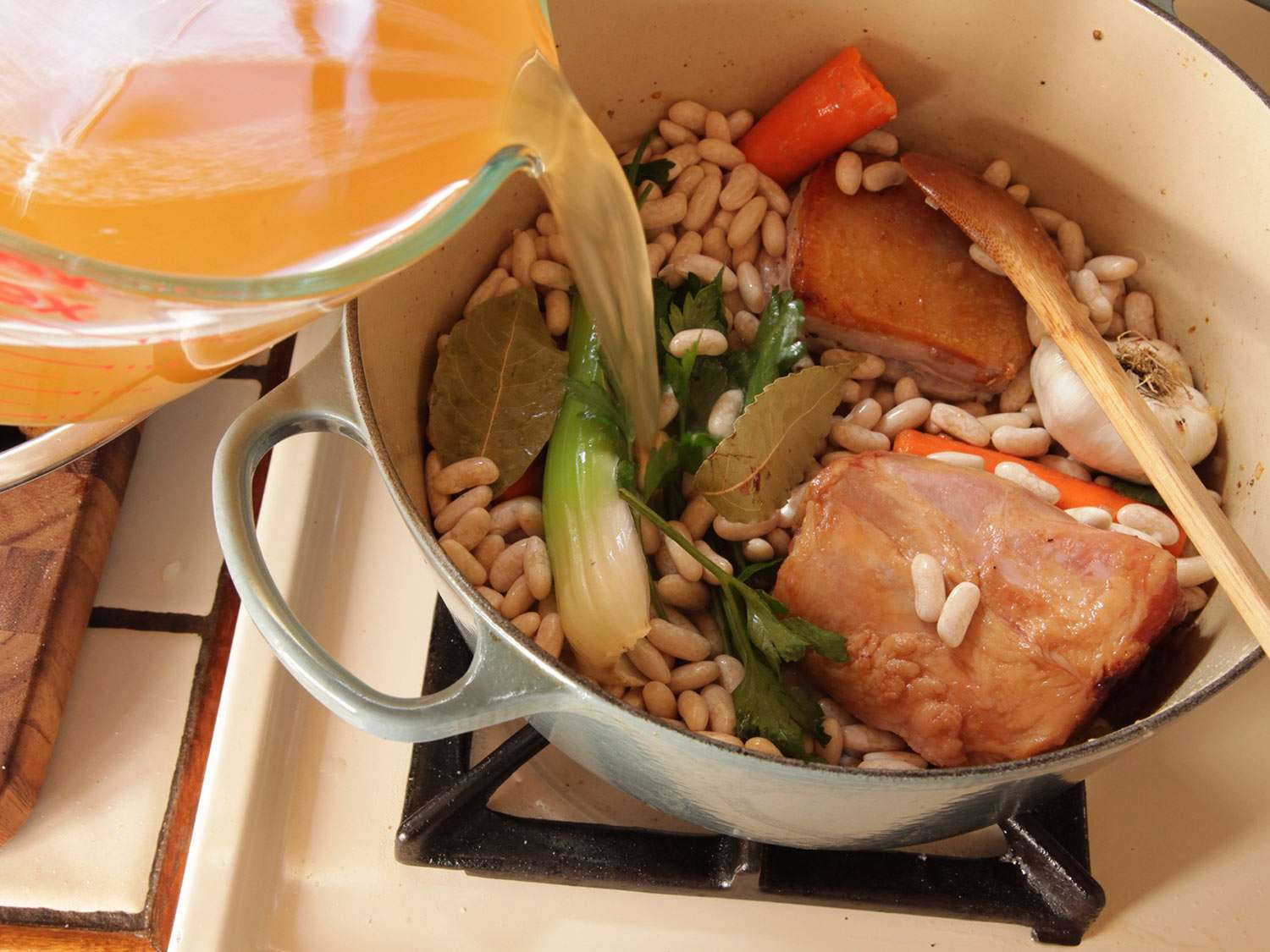 Adding stock to pot of dried beans, salt pork, and aromatics for cassoulet.