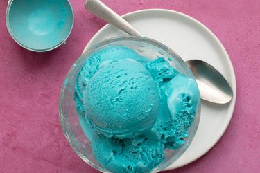 蓝月冰淇淋装在一个玻璃碗里，放在一个白色的陶瓷盘子里，盘子里还有一个金属勺子。背景是粉红色的，图像的左上角是冰淇淋勺，用来舀冰淇淋。