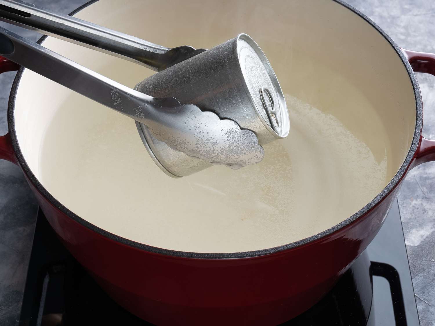 Can of dulce de leche being removed from a cast iron pan of simmering water with metal tongs