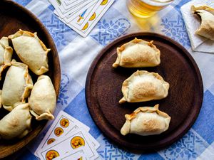 桌子场景的俯视图。两盘肉馅卷饼放在传统的阿根廷蓝白方格织物上。在画框的中间是一张黑色的木桌，上面摆着3个肉馅卷饼排成一排，周围是一瓶啤酒、几张扑克牌和一个吃了一半的肉馅卷饼。